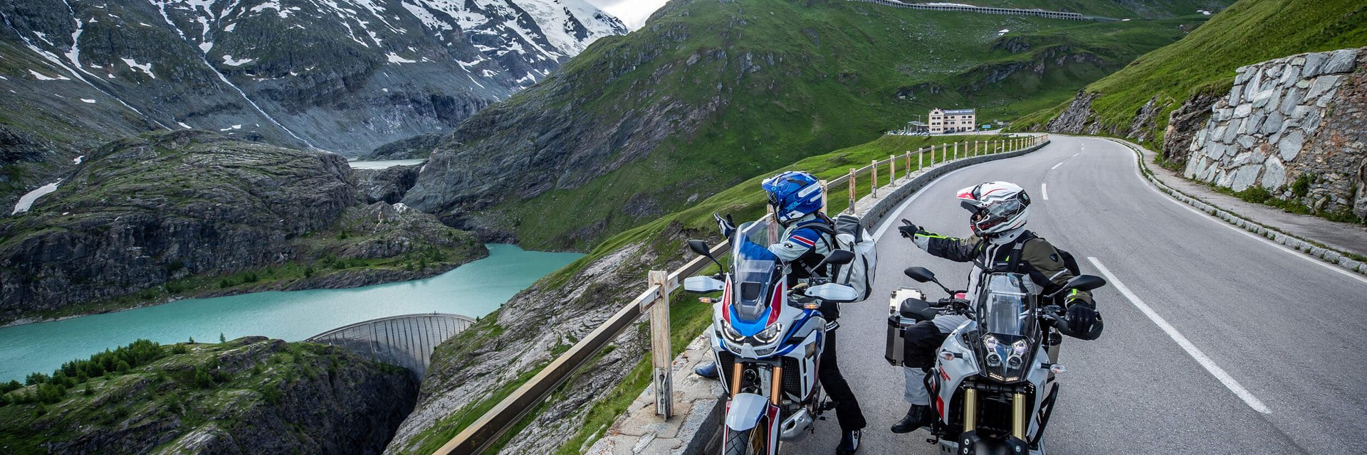 Zwei Motorradfahrer auf Alpenstraße mit Bergsee und schneebedeckten Gipfeln