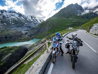 Zwei Motorradfahrer auf Alpenstraße mit Bergsee und schneebedeckten Gipfeln