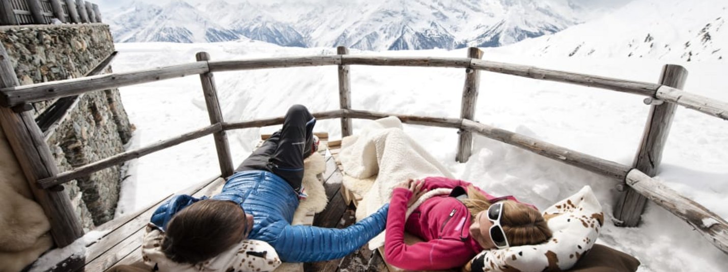 Zwei Personen entspannen auf einer Holzterrasse mit Blick auf verschneite Berge