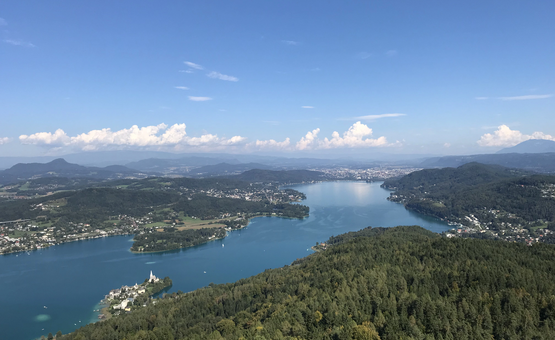 Blick auf einen See mit umliegenden Wäldern und Bergen unter blauem Himmel