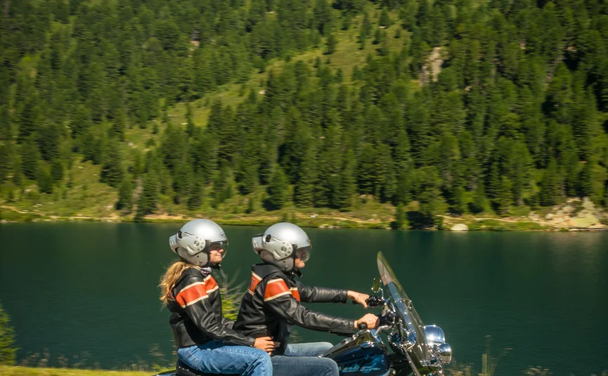 Two people riding a motorcycle near a lake with mountains in the background