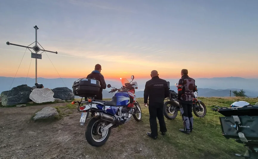 Motorbike holidays in the Wolfsberg holiday region - Lavanttal region © Viktor Sator Three motorcyclists watch sunset from mountain peak with summit cross