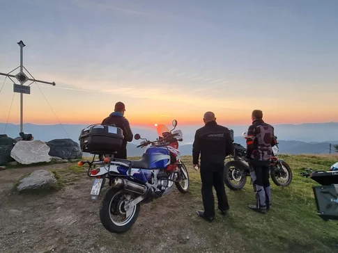 Drei Motorradfahrer schauen bei Sonnenuntergang vom Berggipfel mit Gipfelkreuz