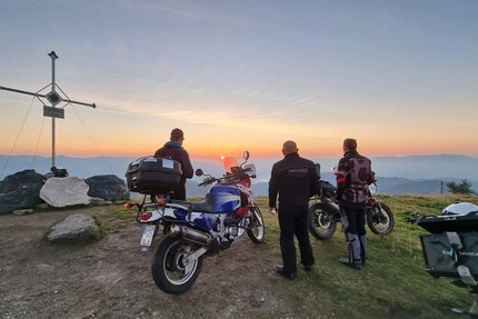 Drei Motorradfahrer schauen bei Sonnenuntergang vom Berggipfel mit Gipfelkreuz