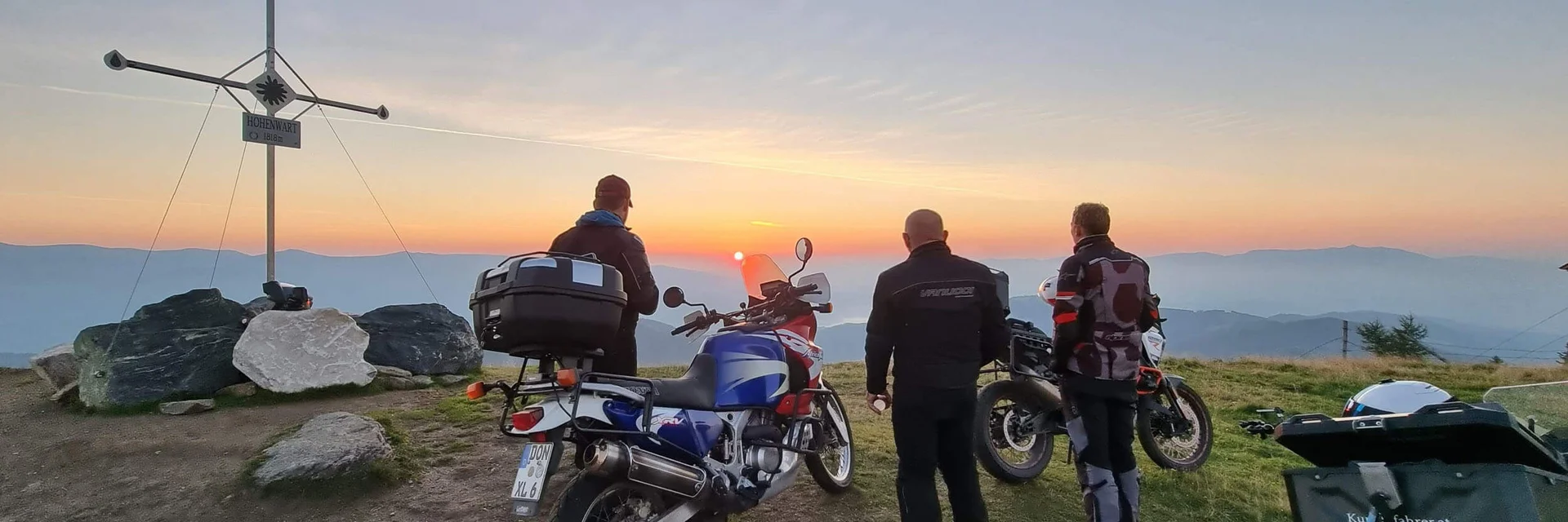 Three motorcyclists watch sunset from mountain peak with summit cross