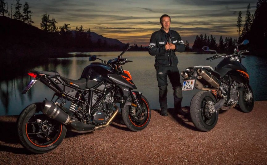 BIKEGENUSS "Großglockner" Person steht zwischen zwei Motorrädern an einem See bei Sonnenuntergang