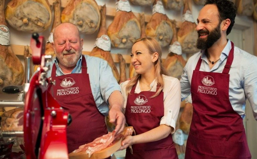 Drei Verkäufer im Prosciutto-Geschäft mit roten Schürzen, die Schinken probieren.