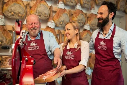 Three shop workers in red aprons tasting prosciutto in a ham store.
