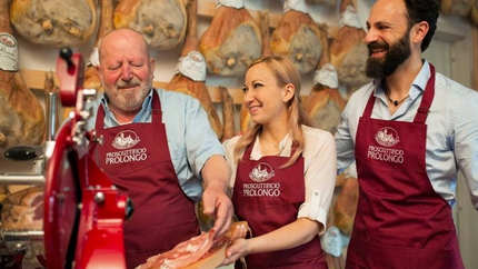 Drei Verkäufer im Prosciutto-Geschäft mit roten Schürzen, die Schinken probieren.