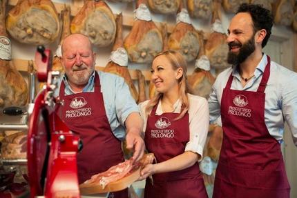 Drei Verkäufer im Prosciutto-Geschäft mit roten Schürzen, die Schinken probieren.