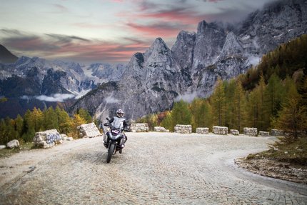 Motorradfahrer auf kurviger Bergstraße vor Felsgipfeln und herbstlichen Bäumen