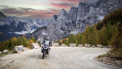 Motorcoureur op kronkelende bergweg met rotsachtige pieken en herfstbomen