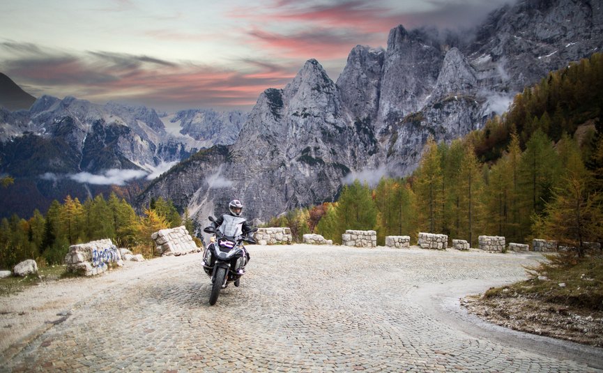 Motorradfahrer auf kurviger Bergstraße vor Felsgipfeln und herbstlichen Bäumen