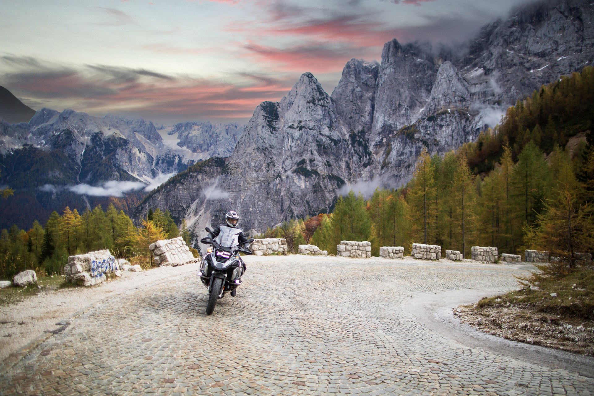 Motorradfahrer auf kurviger Bergstraße vor Felsgipfeln und herbstlichen Bäumen