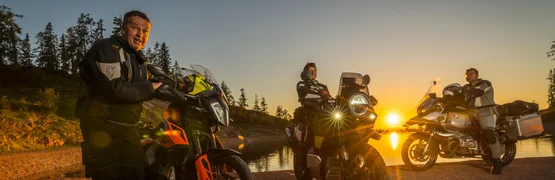 Drei Motorradfahrer stehen am Seeufer bei Sonnenuntergang