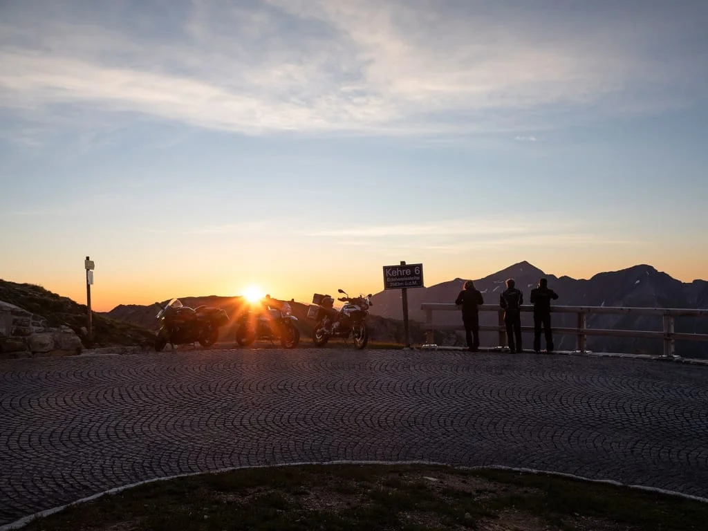 Motorcycles parked at sunset on mountain road with three people by railing