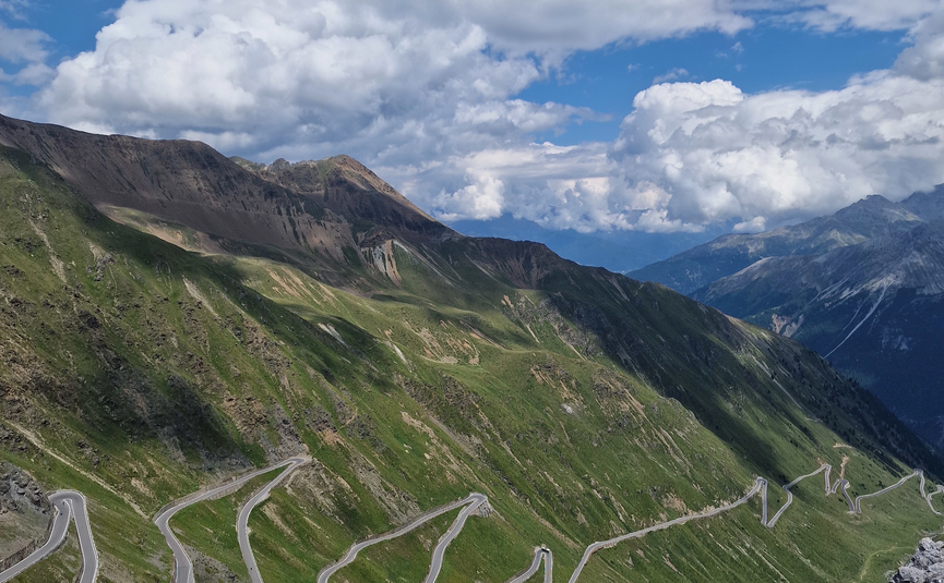 MoHo Schönauer Hof Tour Stelvio Variant 3 Livigno Winding mountain road with sharp turns on a green hillside under a cloudy blue sky