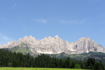Sonnleiten 16: Wilder-Kaiser-Runde („Koasaumrundung“) Felsige Bergkette unter klarem blauen Himmel mit Wald im Vordergrund
