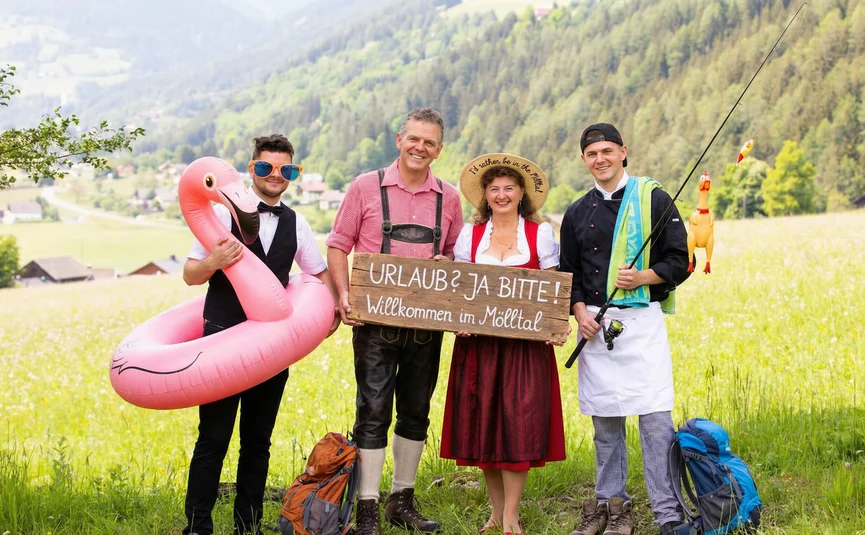 Vier Personen mit Schild im Mölltal, mit aufblasbarem Flamingo und Angelrute auf einer Bergwiese