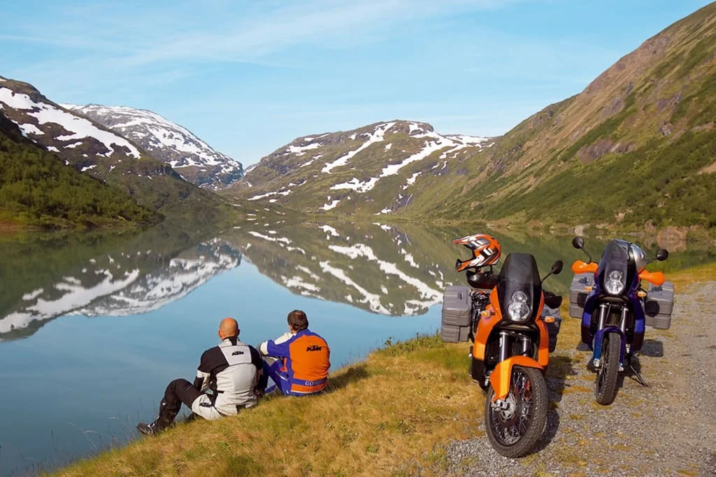 Zwei Motorradfahrer sitzen am See vor schneebedeckten Bergen und reflektiertem Himmel