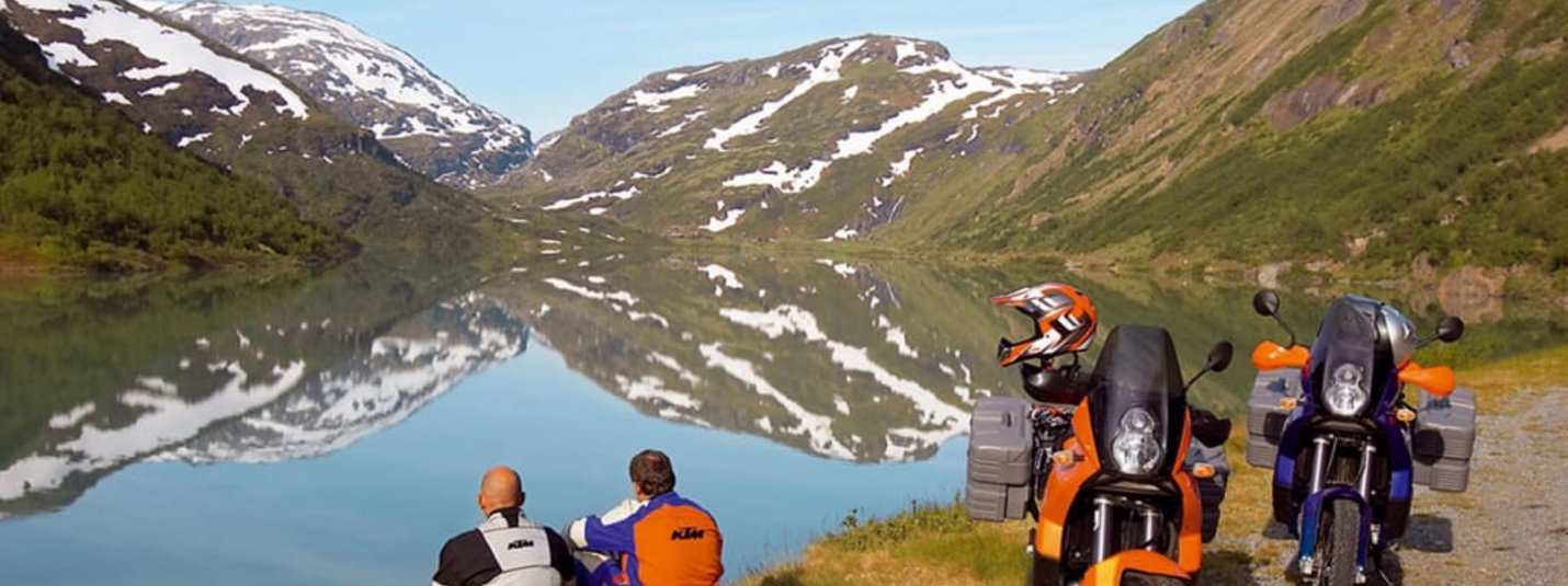 Motorradkurztripp Zwei Motorradfahrer sitzen am See vor schneebedeckten Bergen und reflektiertem Himmel