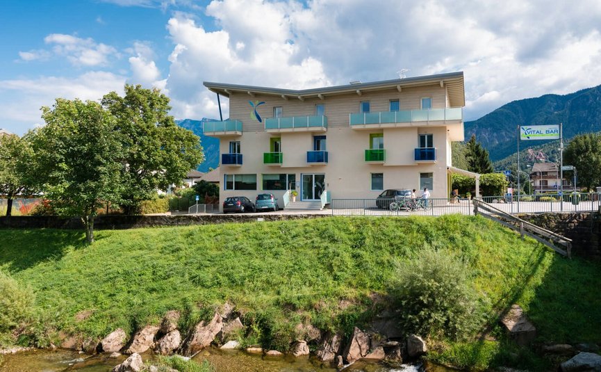 Hotelgebäude mit bunten Balkonen an einem Flussufer vor Bergkulisse