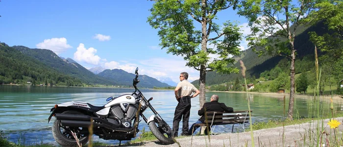 Motorcycle by lake with two men, mountains, and blue sky in background