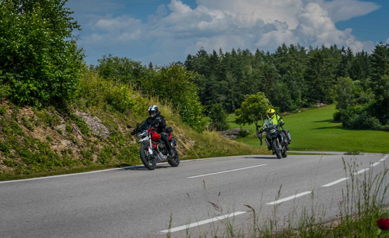 Zwei Motorradfahrer auf einer kurvigen Landstraße mit grünen Hügeln und Wald