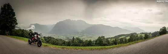 Motorrad auf Landstraße vor Berglandschaft und Tal unter bewölktem Himmel