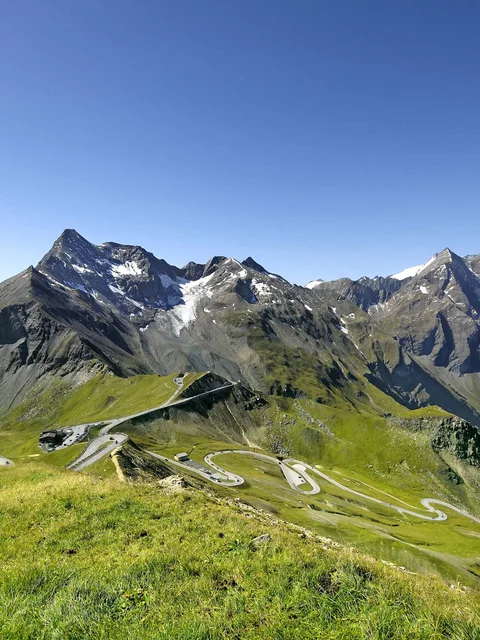 Motorradurlaub © Großglockner Hochalpenstraßen AG Serpentinenstraße in grünen Alpen mit schneebedeckten Bergen unter blauem Himmel