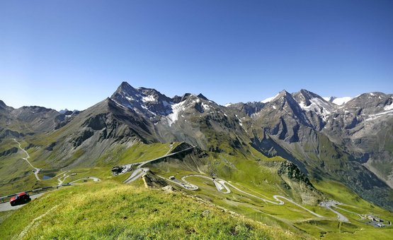 Kronkelende bergweg in groene Alpen met besneeuwde toppen onder een heldere blauwe lucht