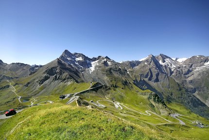 Kronkelende bergweg in groene Alpen met besneeuwde toppen onder een heldere blauwe lucht