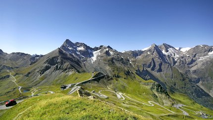 Kronkelende bergweg in groene Alpen met besneeuwde toppen onder een heldere blauwe lucht