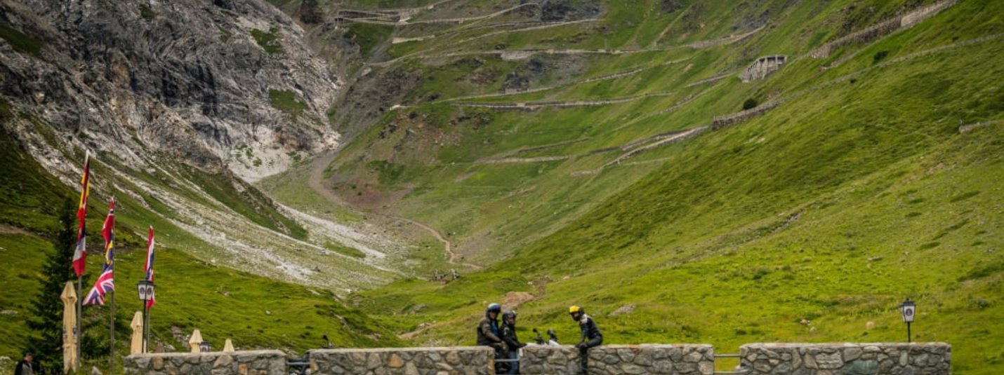 Steinbrücke mit Motorradfahrern vor grünen Bergen und Serpentinenstraße