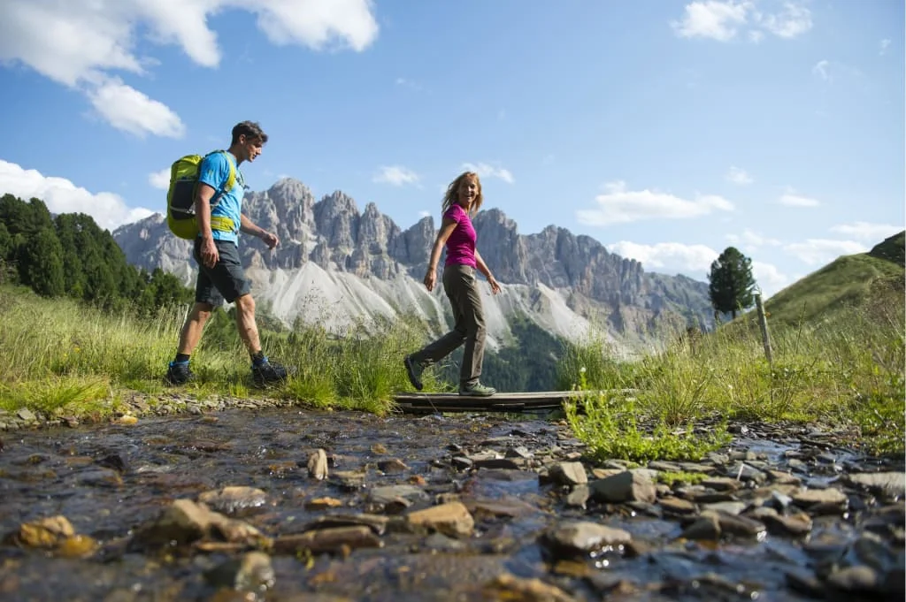 Two hikers crossing a stream with mountain scenery in the background