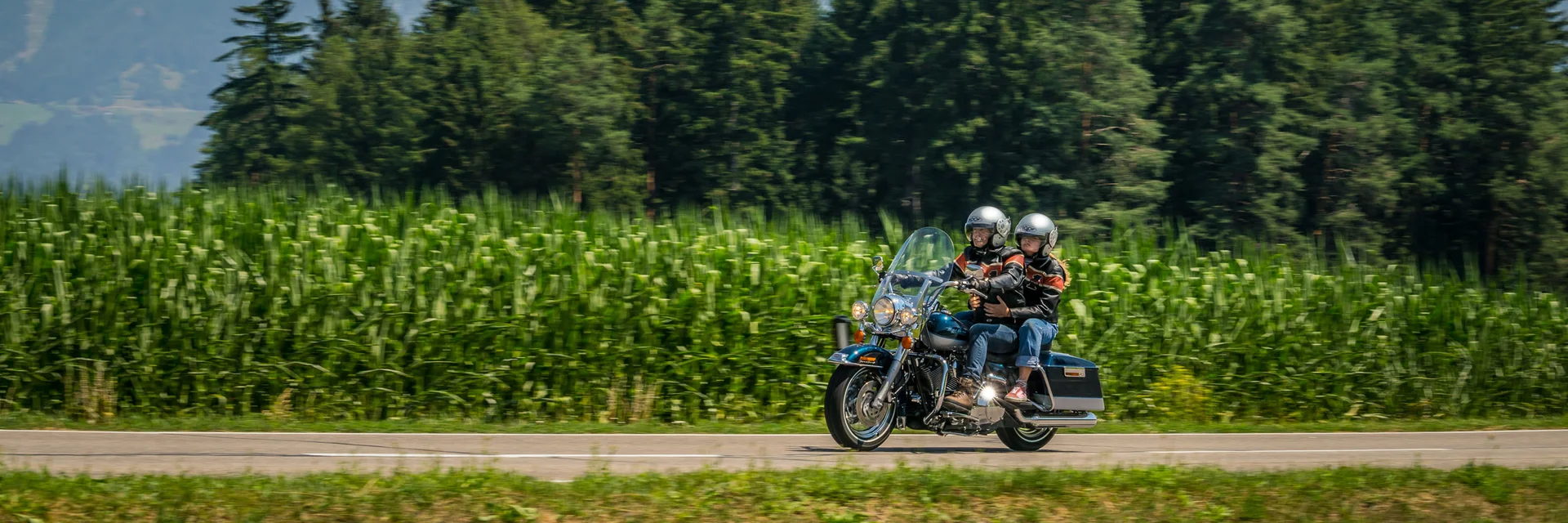 Two motorcyclists riding on a road with forest and mountains background