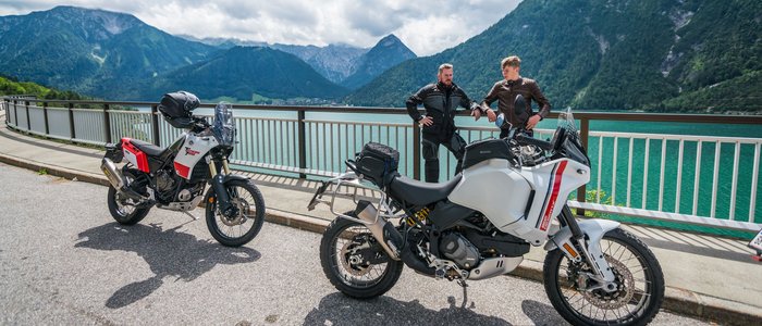 Zwei Männer mit Motorrädern vor Bergsee und Bergen bei bewölktem Himmel