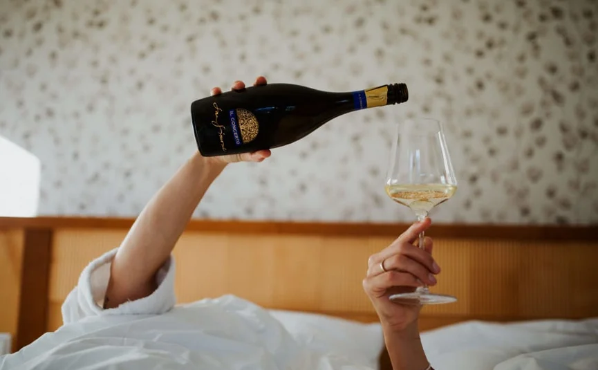Person gießt Weißwein aus Flasche in Weinglas im Bett