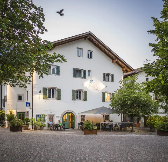 Tour 1 Dolomitenpässe - Gröden - Regglberg Km 350 © Hannes Niederkofler Café mit Außensitzplätzen an einem kopfsteingepflasterten Platz umgeben von Bäumen