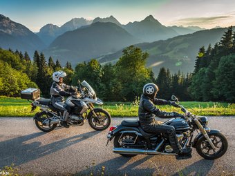 Zwei Motorradfahrer auf Bergstraße mit Alpenkulisse bei Sonnenuntergang