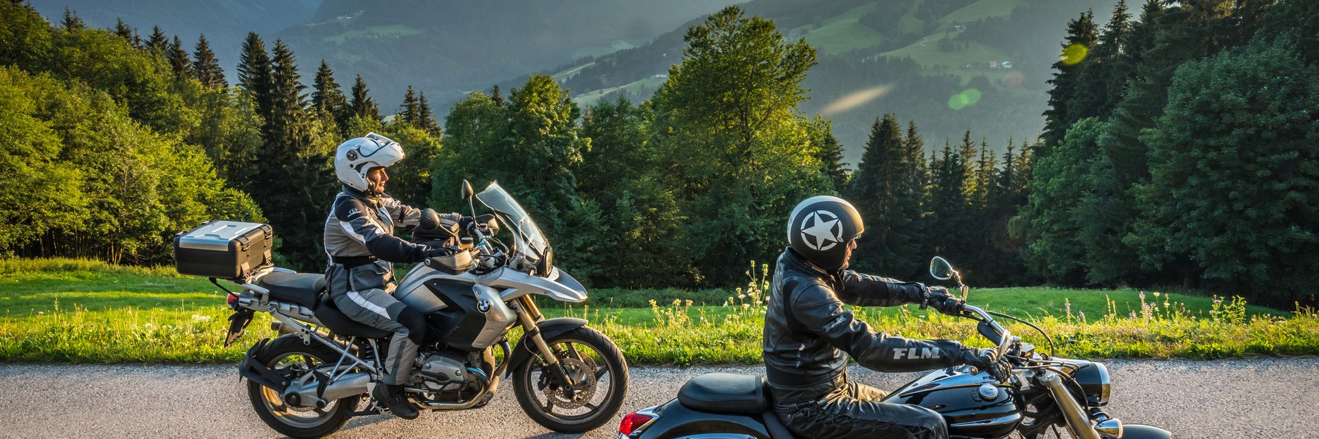 Zwei Motorradfahrer auf Bergstraße mit Alpenkulisse bei Sonnenuntergang