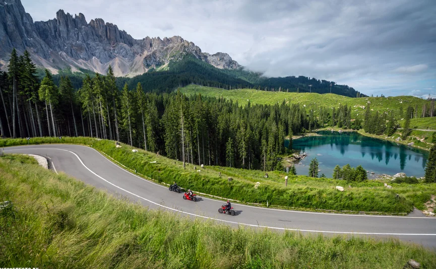Bike & Ski Hotel Diana Karersee ***S © Moppetfoto.de Motorcyclists on a mountain road beside a turquoise lake, surrounded by forest and rocky peaks