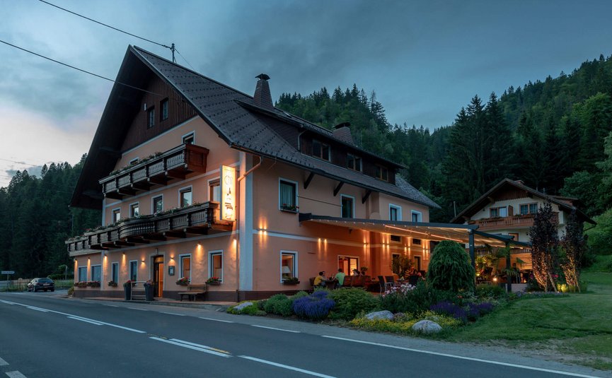 Beleuchtetes Gasthaus am Straßenrand in bewaldeter Gebirgslandschaft bei Abenddämmerung