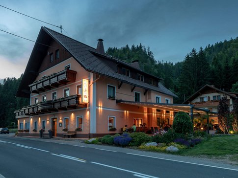 Beleuchtetes Gasthaus am Straßenrand in bewaldeter Gebirgslandschaft bei Abenddämmerung