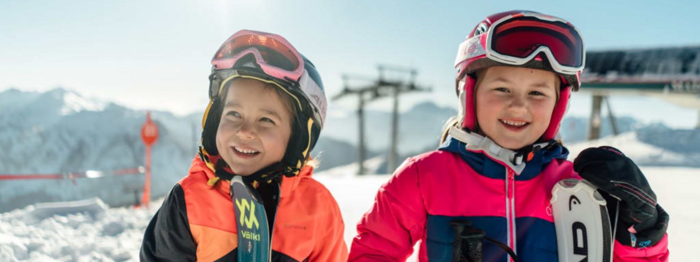 Leistbarer Familienskiurlaub Zwei lachende Kinder mit Skihelmen und Skiern im Schnee bei Sonnenschein