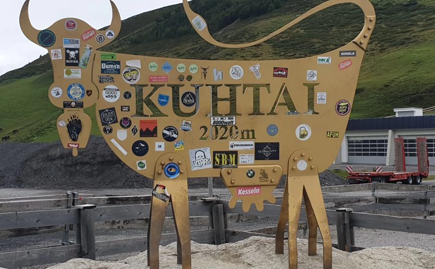 Große goldene Kuh-Figur mit 'KUHTAI 2020m' und vielen Aufklebern in den Bergen.