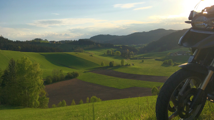 Motorrad im Vordergrund vor grünen Hügeln und Tal bei Sonnenuntergang