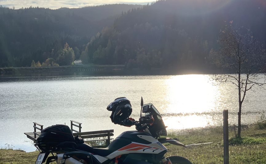 Motorrad am Seeufer bei Sonnenuntergang mit bewaldeten Hügeln im Hintergrund