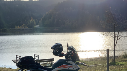 Motorrad am Seeufer bei Sonnenuntergang mit bewaldeten Hügeln im Hintergrund