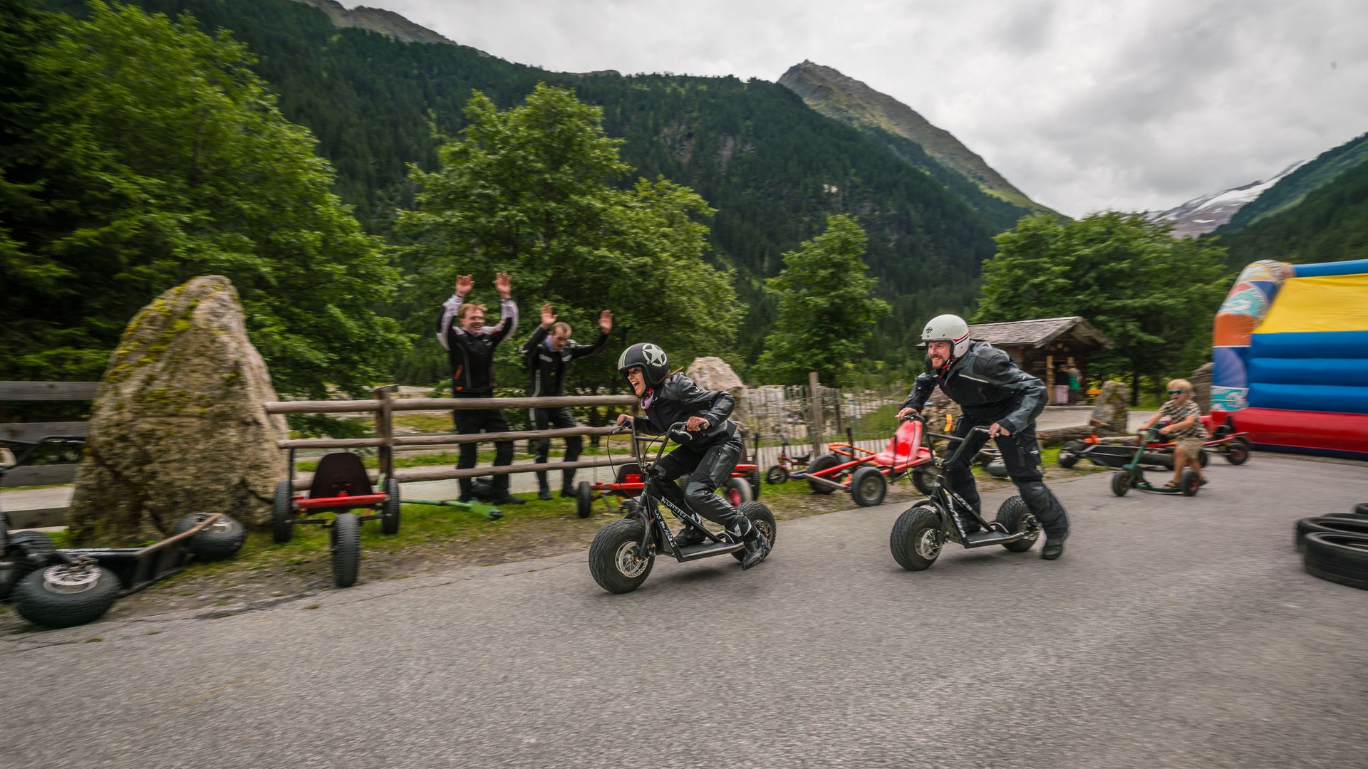 Zwei Personen fahren auf motorisierten Tretrollern bergauf, Zuschauer jubeln